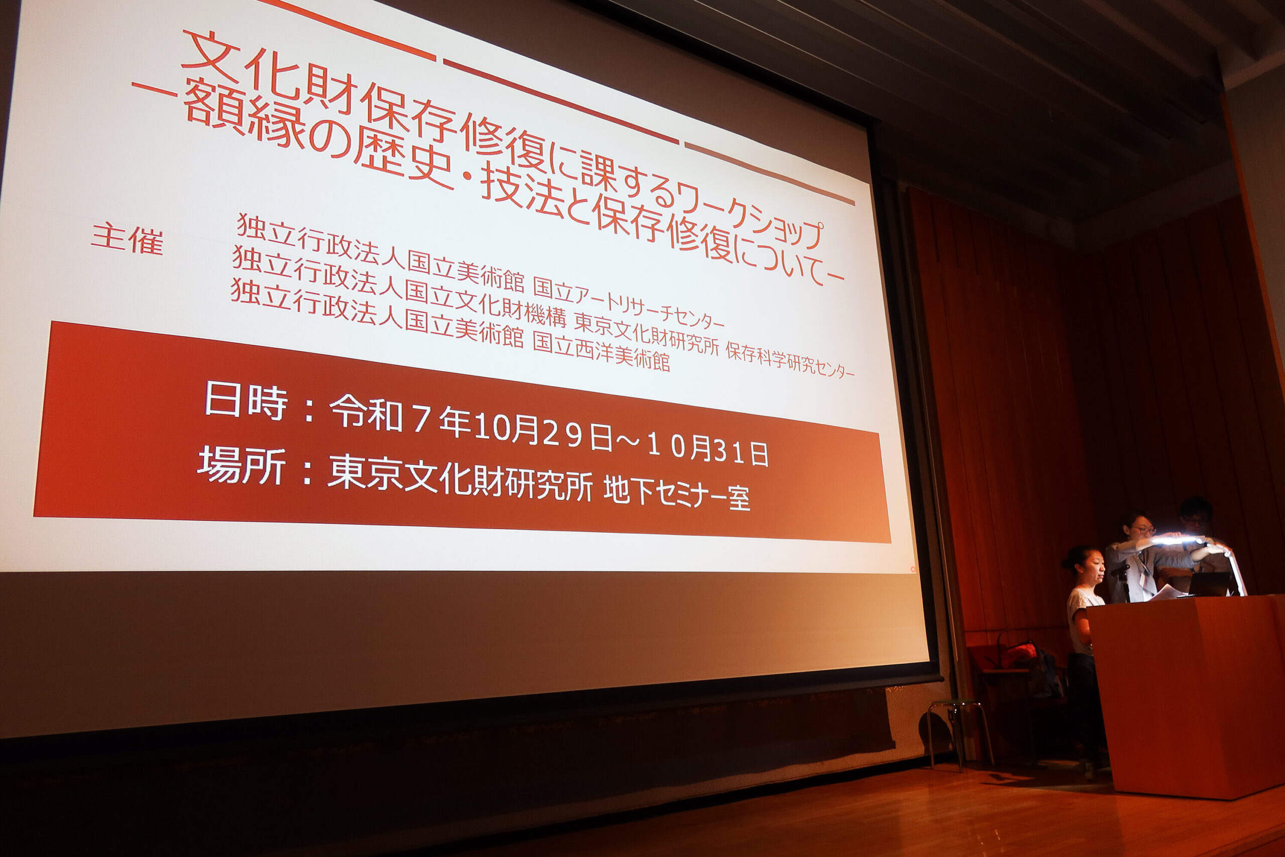 この写真には、講演会の様子（講演会のタイトルと日時が画面に投影されている）が写されています。