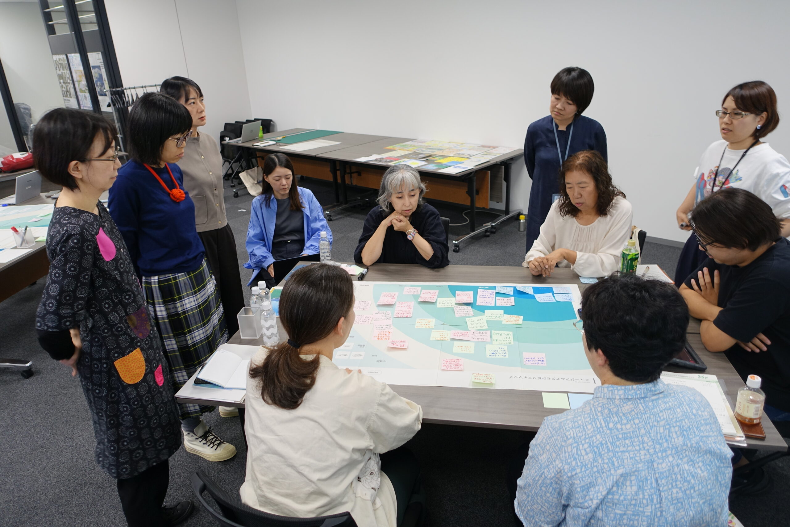 11名の人物が、一つのテーブルを囲って話し合っている。テーブルの上には大きな紙が広がっており、数々のふせんが貼られている。