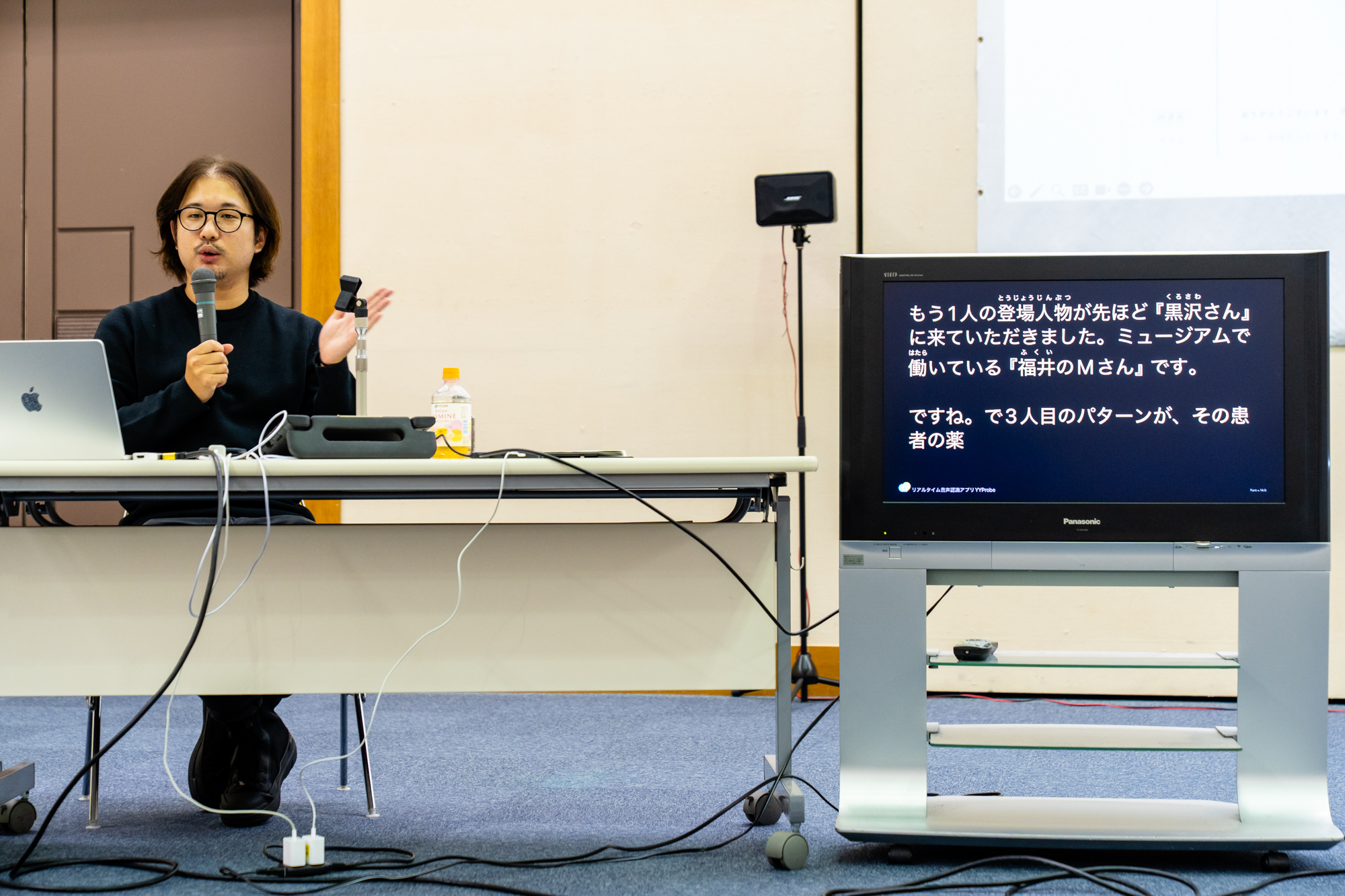 マイクを持った人物が話しをしている様子。右側には黒いモニターがあり、音声認識アプリケーションによって話した言葉が白い文字で表示されている。