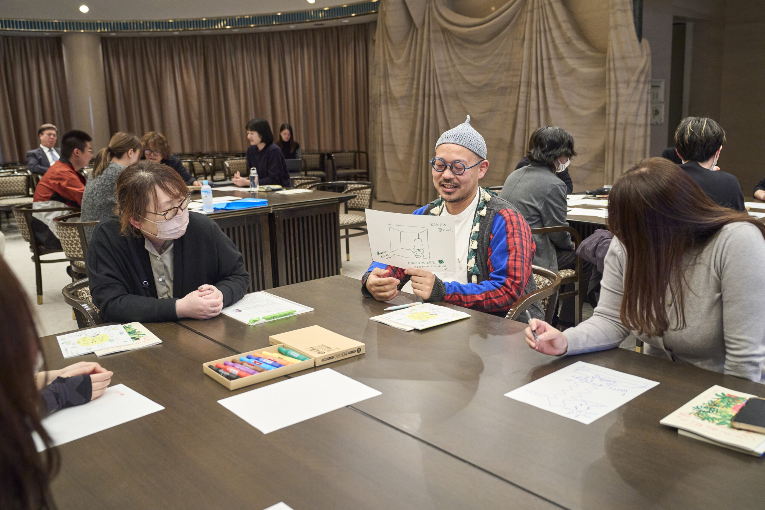 3人の人物がテーブルに座っている。中央の人物が、白い紙に書かれたイラストを持ち笑顔で話しており、両側の2人がのぞきこんでいる。